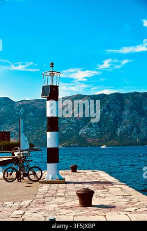 A vertical daytime composition showcasing a small lighthouse with black and white stripes standing on a stone pier, accompanied by a parked bicycle, with the blue sea and massive mountains in the background. Stock Photo