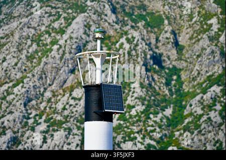 A close-up photograph focusing on the top section of a small black and white striped navigational lighthouse, equipped with a solar panel, safety rail, and beacon light, set against a backdrop of a rugged, textured mountain slope. Stock Photo
