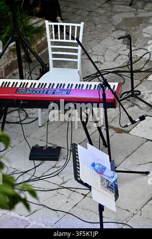 A high-angle shot of a music performance area on a stone terrace. A red stage keyboard is positioned in front of a white Chiavari chair. Multiple microphone stands, cables, and a music stand with papers and a water bottle are set up, ready for a musician. Stock Photo