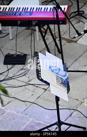 A high-angle shot of a music performance area on a stone terrace. A red stage keyboard is positioned in front of a white Chiavari chair. Multiple microphone stands, cables, and a music stand with papers and a water bottle are set up, ready for a musician. Stock Photo