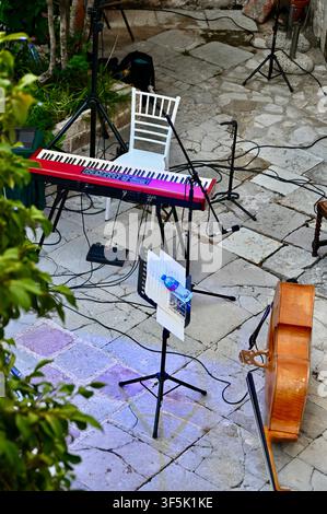 A high-angle shot of a music performance area on a stone terrace. A red stage keyboard is positioned in front of a white Chiavari chair. Multiple microphone stands, cables, and a music stand with papers and a water bottle are set up, ready for a musician. Stock Photo