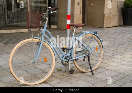 A light blue bicycle with a brown basket is locked to a metal pole on a sidewalk, waiting for its rider to return after finishing business at the near Stock Photo