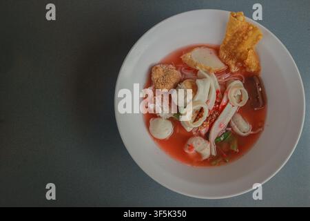 Top view Thai Yentafo pink noodle soup with fish balls, squid, pork blood and crispy wonton in a white bowl on dark background. Stock Photo