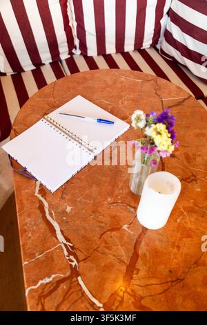 Open spiral notebook with blue pen, colorful flowers, and white candle arranged on a warm marble cafe table beside striped seating, creating a cozy li Stock Photo