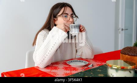 Young woman drinking coffee at a festively decorated dining table during a New Year breakfast at home, with panettone, candle and seasonal table decor Stock Photo