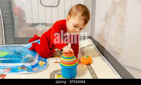 Curious baby in red pajamas plays with colorful stacking cups inside a soft playpen at home, exploring motor skills and early learning through safe pl Stock Photo