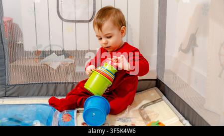 Curious toddler in red pajamas sits inside a safe mesh playpen, stacking colorful plastic cups on a soft mat at home, practicing fine motor skills and Stock Photo