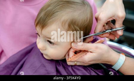Close-up of a toddler getting a haircut at a salon, with stylist hands using scissors and comb near the ear, wearing a protective cape, calm and focus Stock Photo