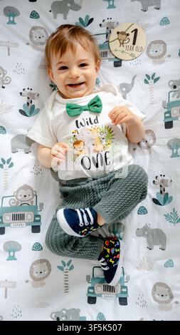 Smiling toddler boy lying on safari themed blanket, wearing white wild one shirt with green bow tie and holding 1 year old milestone card for first bi Stock Photo