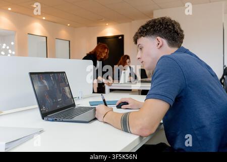 MBA student attends an online class on a laptop while taking notes in a modern business school workspace, with fellow students collaborating at desks Stock Photo