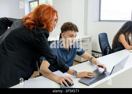 Professor provides one-on-one guidance to an MBA student using a laptop in a bright classroom, while another student studies nearby during a business Stock Photo