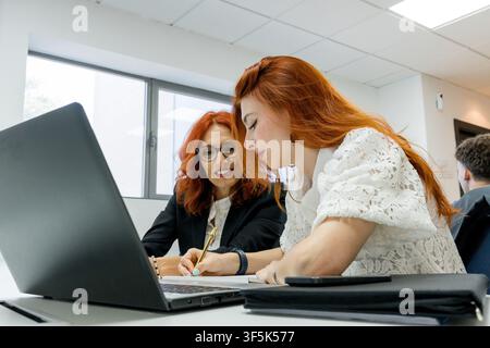 Two MBA students collaborate on a laptop assignment in a modern classroom, reviewing notes and writing together during a business education session. Stock Photo