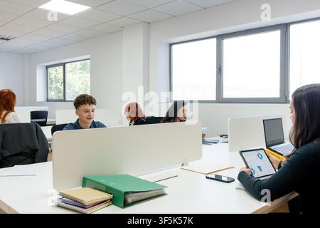 MBA students work independently and collaborate at desks in a bright modern university classroom, using laptops, tablets, notebooks, and study materia Stock Photo