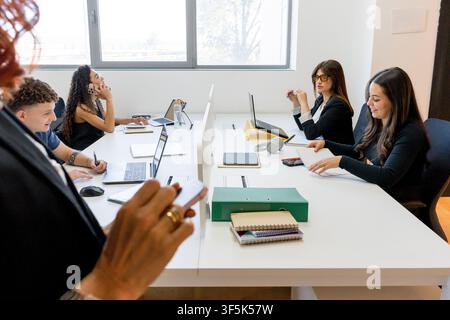 MBA students collaborate at a shared desk in a modern classroom, using laptops, notebooks and smartphones during a business workshop and group study s Stock Photo