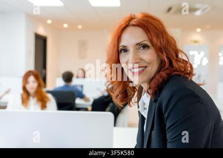Smiling MBA mentor in a modern business classroom looks at camera while students work on laptops in the background, representing leadership, education Stock Photo