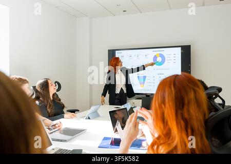 MBA students listen as an instructor presents business analytics charts on a digital screen during a collaborative classroom lecture with laptops and Stock Photo