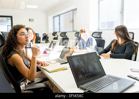 MBA students collaborate around a conference table in a bright university classroom, using laptops and notes during a business discussion, team projec Stock Photo