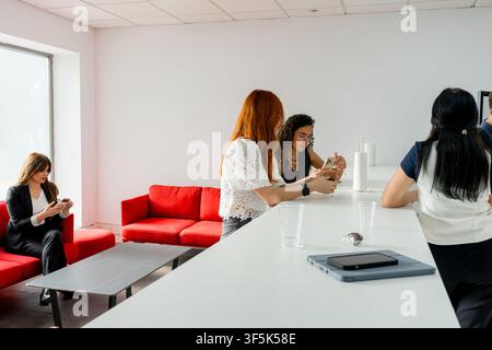 MBA students relax during a break in a modern business school lounge, using smartphones and talking beside a white counter with red sofas and a tablet Stock Photo