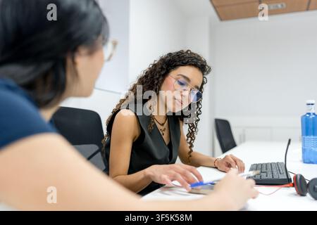 Two MBA students collaborate on coursework at a desk in a modern university classroom, reviewing documents and using a desktop computer during a focus Stock Photo