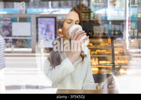 Young businesswoman enjoys takeaway coffee while using a smartphone outside a bakery cafe, seen through a reflective shop window during a work break. Stock Photo