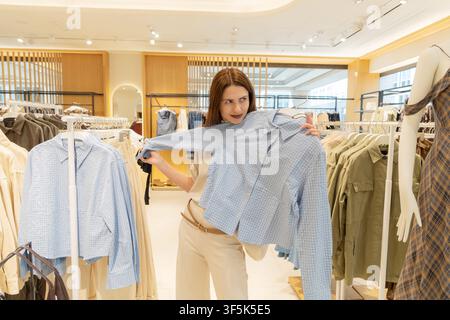 Smiling woman compares a checked blouse while shopping in a bright fashion clothing store, surrounded by garment racks, casual apparel and a mannequin Stock Photo