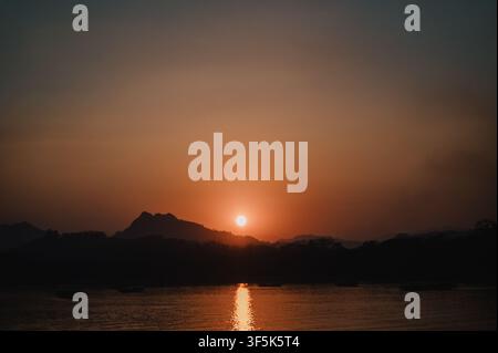 Golden sunset illuminates the Mekong River in Laos, with a glowing sun reflected on calm water and dark mountain silhouettes beneath a dramatic evenin Stock Photo