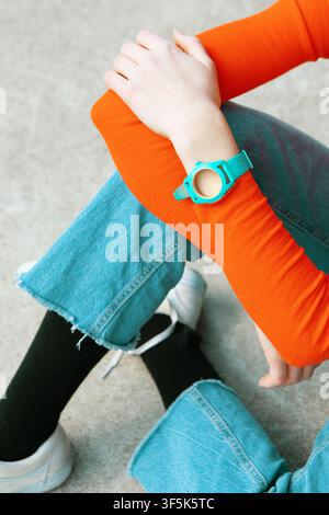 Top view of a person seated on concrete, wearing a bright orange sleeve, turquoise wristwatch, cropped denim jeans, black socks, and white sneakers. Stock Photo