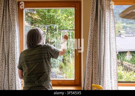 Mature man cleans a large window with a squeegee in a cozy apartment, seen from behind beside patterned curtains and overlooking a leafy residential n Stock Photo