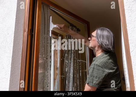 Mature man cleans a large apartment window with a squeegee, removing water and dirt from the glass indoors during routine household maintenance. Stock Photo