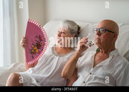 Senior couple rest indoors during extreme summer heat, using a hand fan and drinking water to prevent heat exhaustion and manage climate stress at hom Stock Photo