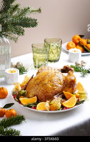 Golden roasted turkey on a white platter with orange slices, kumquat, rosemary, pomegranate, and physalis, framed by evergreen branches, candles, glas Stock Photo