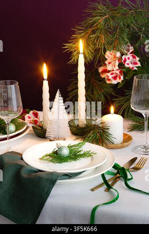 Elegant Christmas table setting with glowing candles, evergreen branches, floral accents, white plates, green napkin, glassware and a glittering ornam Stock Photo