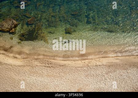 Overhead view of clear Mediterranean water gently reaching a sandy beach in Sicily, Italy. Sunlit pebbles, submerged rocks, and rippled shallows creat Stock Photo