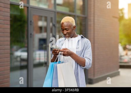 Smiling african american guy with packages with purchases in hands, typing on smartphone Stock Photo