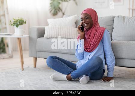 Joyful black islamic lady talking on cellphone, relaxing on floor at home Stock Photo