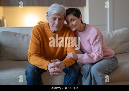 Elderly couple sitting on couch together embracing and supporting each other during difficult time. Stock Photo