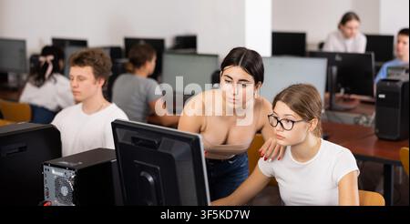 Focused female teenager helping to the positive classmate while she is learning computer science in the class Stock Photo