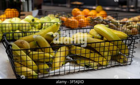 Fresh ripe bananas displayed in a black wire basket at a supermarket produce section with apples and oranges in the background. Healthy grocery shoppi Stock Photo