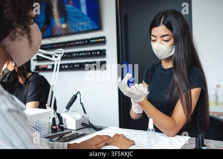 Health care, manicure and social distance. Asian young woman in protective mask wearing rubber gloves for working with nails of african american lady Stock Photo