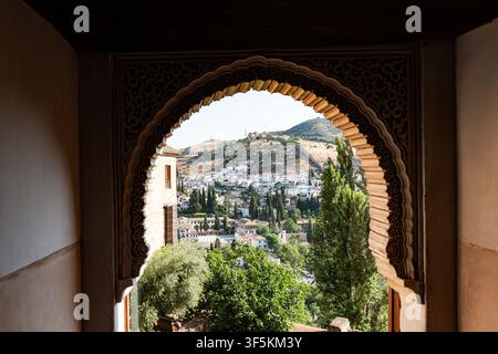 The image shows an ornate, intricately carved Moorish horseshoe arch framing a sunny outdoor view of the historic Albaicín white-washed neighborhood a Stock Photo