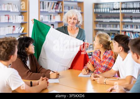 Multiethnic teenager students studying at language class. Mature female educator holding national flag of Italy in library Stock Photo