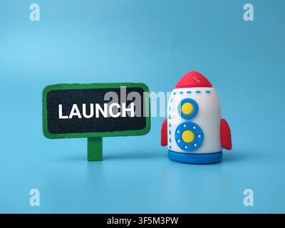 A colorful toy rocket standing beside a small green sign board displaying the word LAUNCH on a blue backdrop. Stock Photo