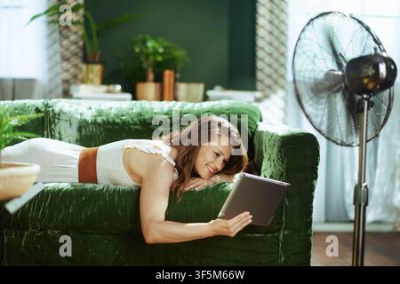 A smiling woman in a stylish white dress & brown belt relaxes on a green velvet sofa, enjoying her digital tablet. The tranquil home setting, with a f Stock Photo