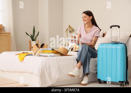 Young woman planning summer vacation trip, writing packing list on bed with Clothes and suitcase Stock Photo