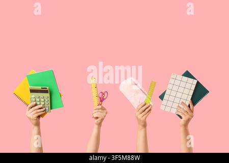 Multiple hands holding various school supplies including notebooks a calculator rulers scissors and a pencil case on a pink background Stock Photo