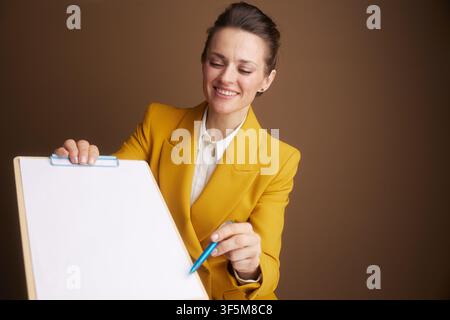 A smiling businesswoman in a yellow blazer looks down at a clipboard with a pen, reviewing information. This image conveys focus, planning, or decisio Stock Photo
