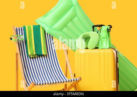Summer vacation travel essentials with yellow suitcase, green inflatable mattress, and striped beach chair on a bright background Stock Photo