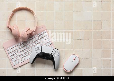 Pink wireless headphones, keyboard, white game controller, and mouse on a beige tiled background, top view Stock Photo