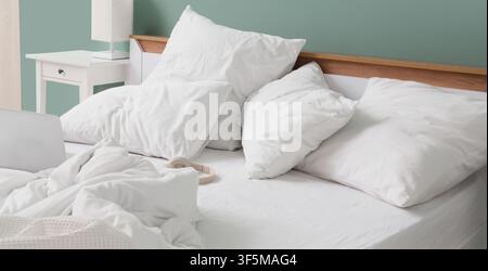 Unmade bed with white linen, pillows, and duvet, a laptop and headphones on the mattress, and a nightstand with a lamp Stock Photo