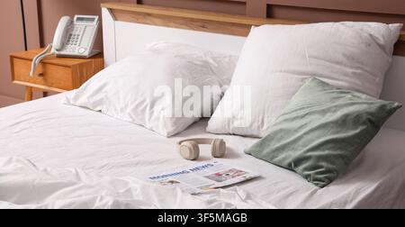 Unmade bed with white sheets, pillows, morning newspaper, headphones, and telephone on a wooden nightstand Stock Photo